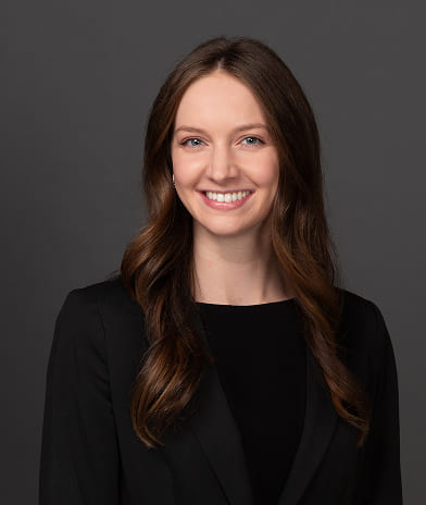 Head-and-shoulders portrait of a smiling woman with long brown hair in a black blazer, against a dark gray background.