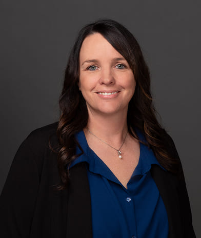 Professional headshot of a smiling woman with dark hair, wearing a blue blouse and black blazer, gray background.