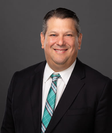 Smiling man in a dark suit and teal plaid tie posing for a professional headshot against a gray background.