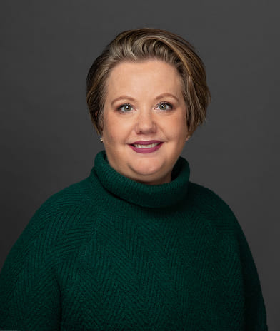Portrait of a smiling woman with short light-brown hair wearing a dark green turtleneck against a gray backdrop and soft lighting.