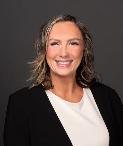 Professional headshot of a smiling woman with shoulder-length light brown hair, wearing a black blazer and white blouse.