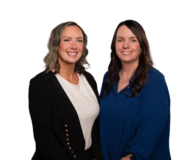 Two women standing side by side, smiling at the camera; one wears a black blazer over a white top, the other a blue blouse.