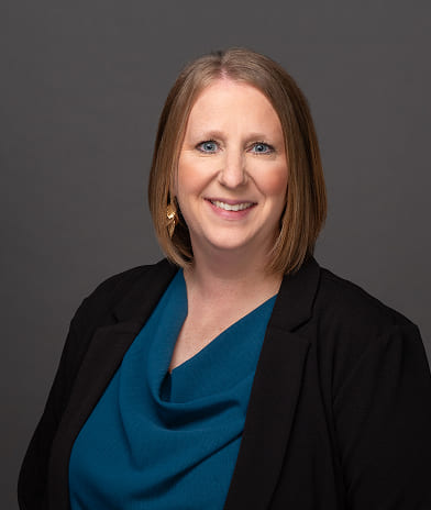 Professional headshot of a smiling woman with shoulder-length brown hair, wearing a teal blouse and black blazer against a gray backdrop.